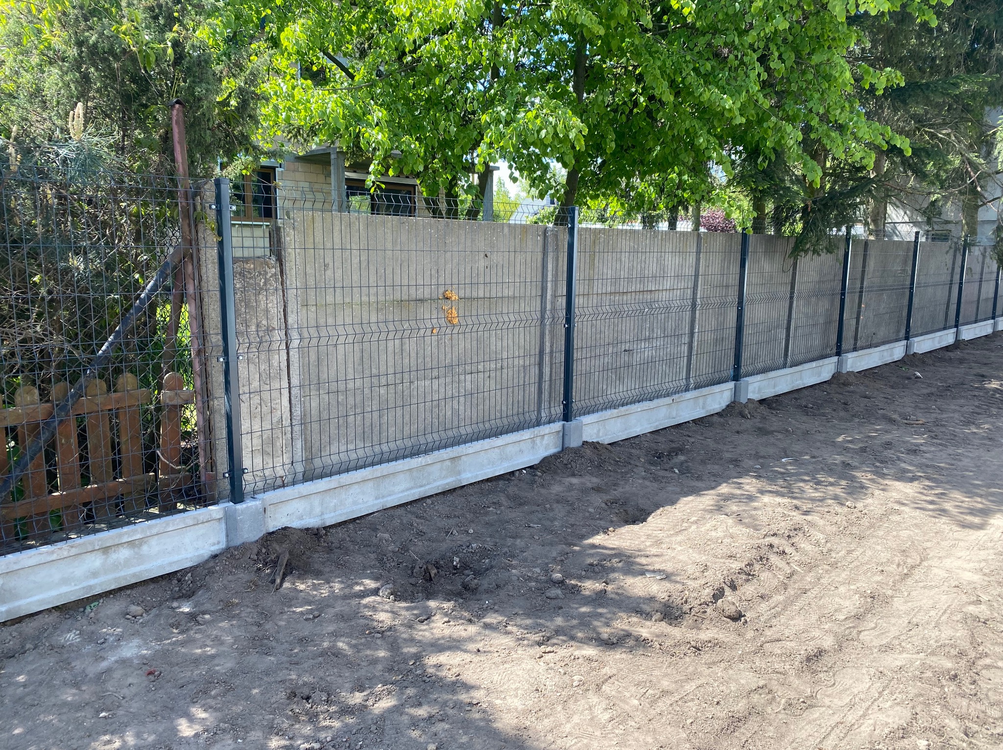 Nowoczesne ogrodzenie panelowe z podmurówką betonową, widok z zewnątrz, zieleń w tle. Szara kolorystyka betonu i paneli kontrastuje z zielenią drzew.