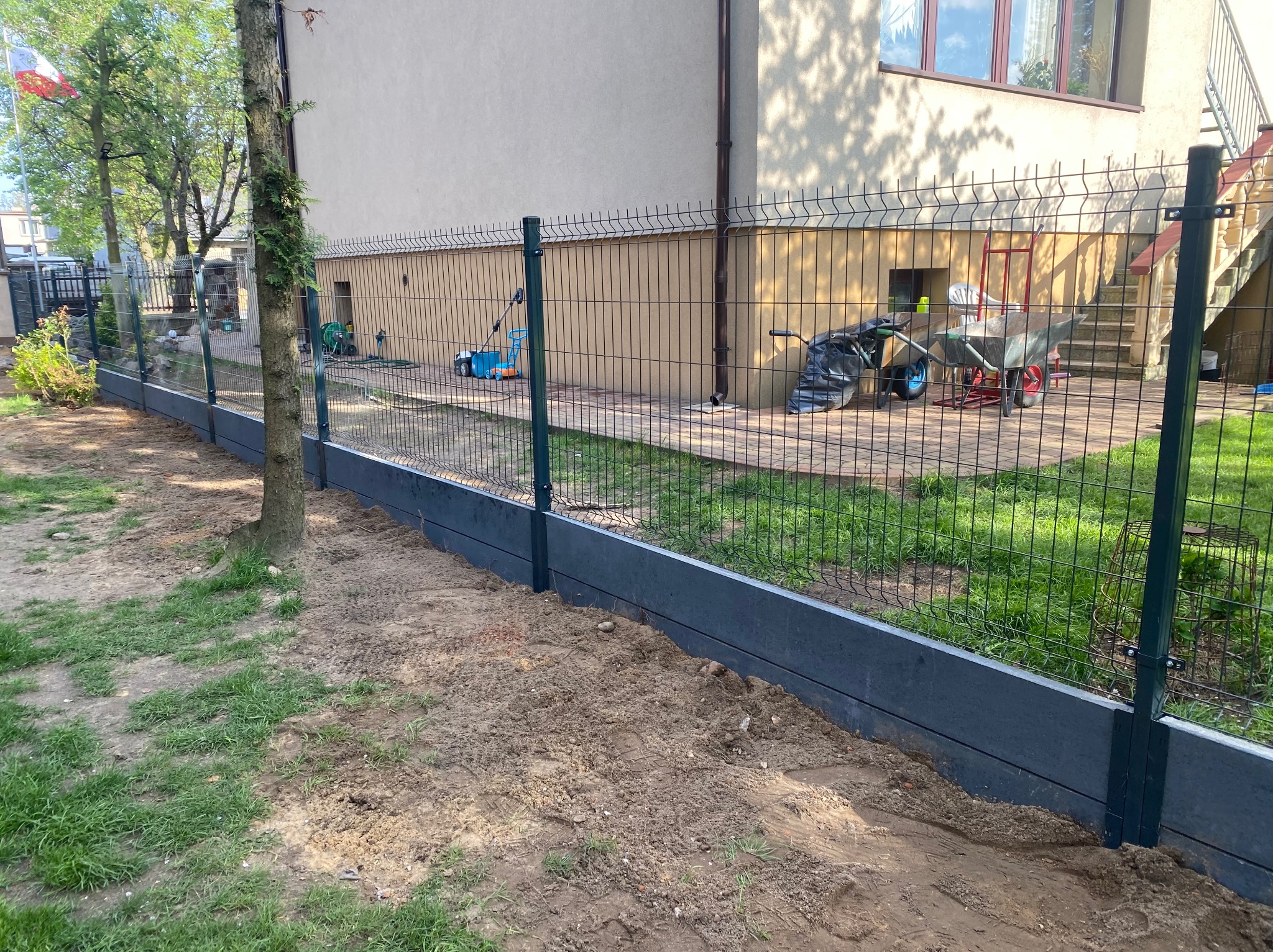 Nowoczesne ogrodzenie panelowe z podmurówką w kolorze antracytowym, widok z bliska na świeżo obsypaną ziemią podstawę. Słupki w kolorze ciemnej zieleni.