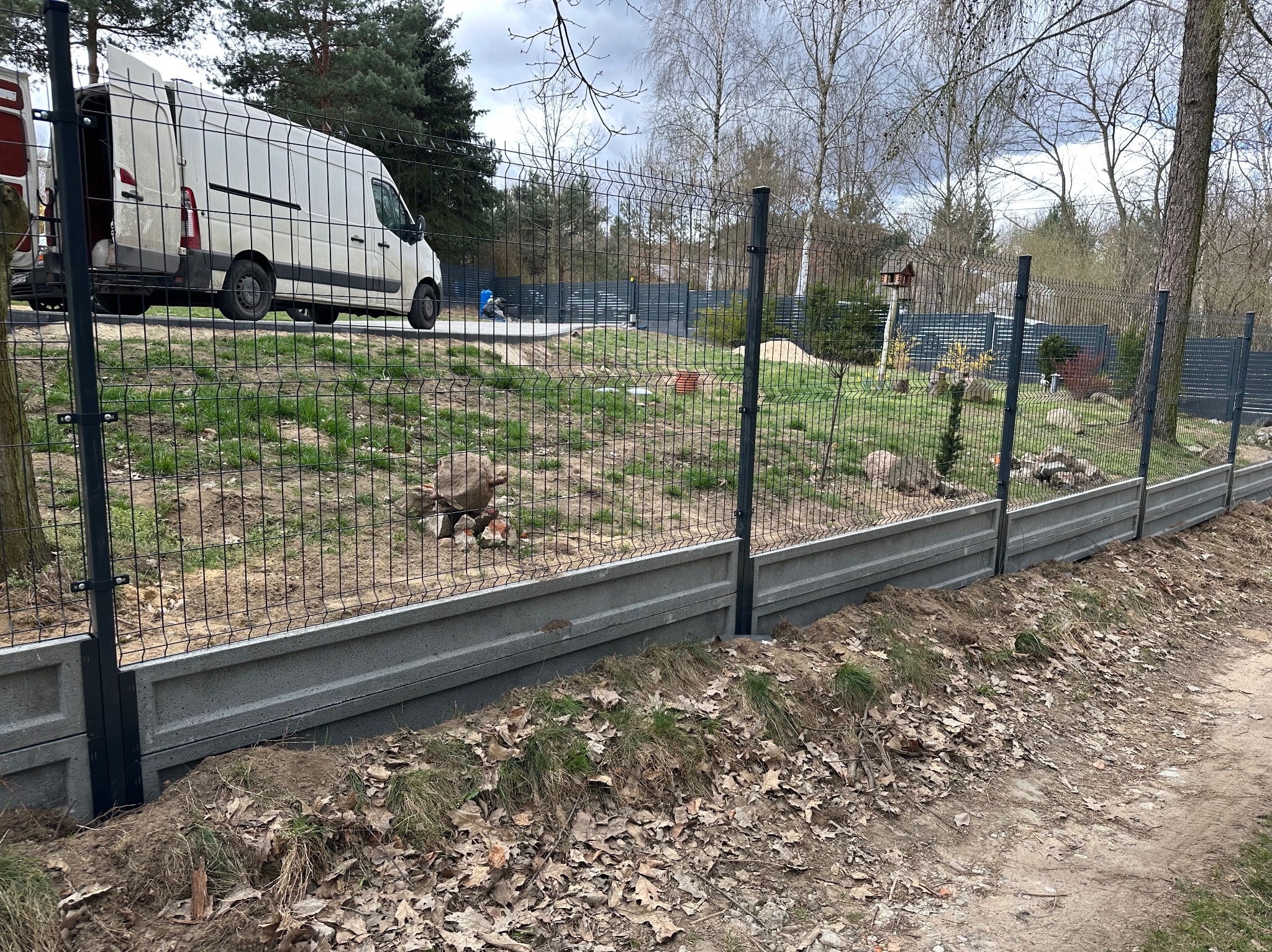 Nowoczesne ogrodzenie panelowe z podmurówką betonową, widok z zewnątrz posesji. Na drugim planie zaparkowany biały bus.