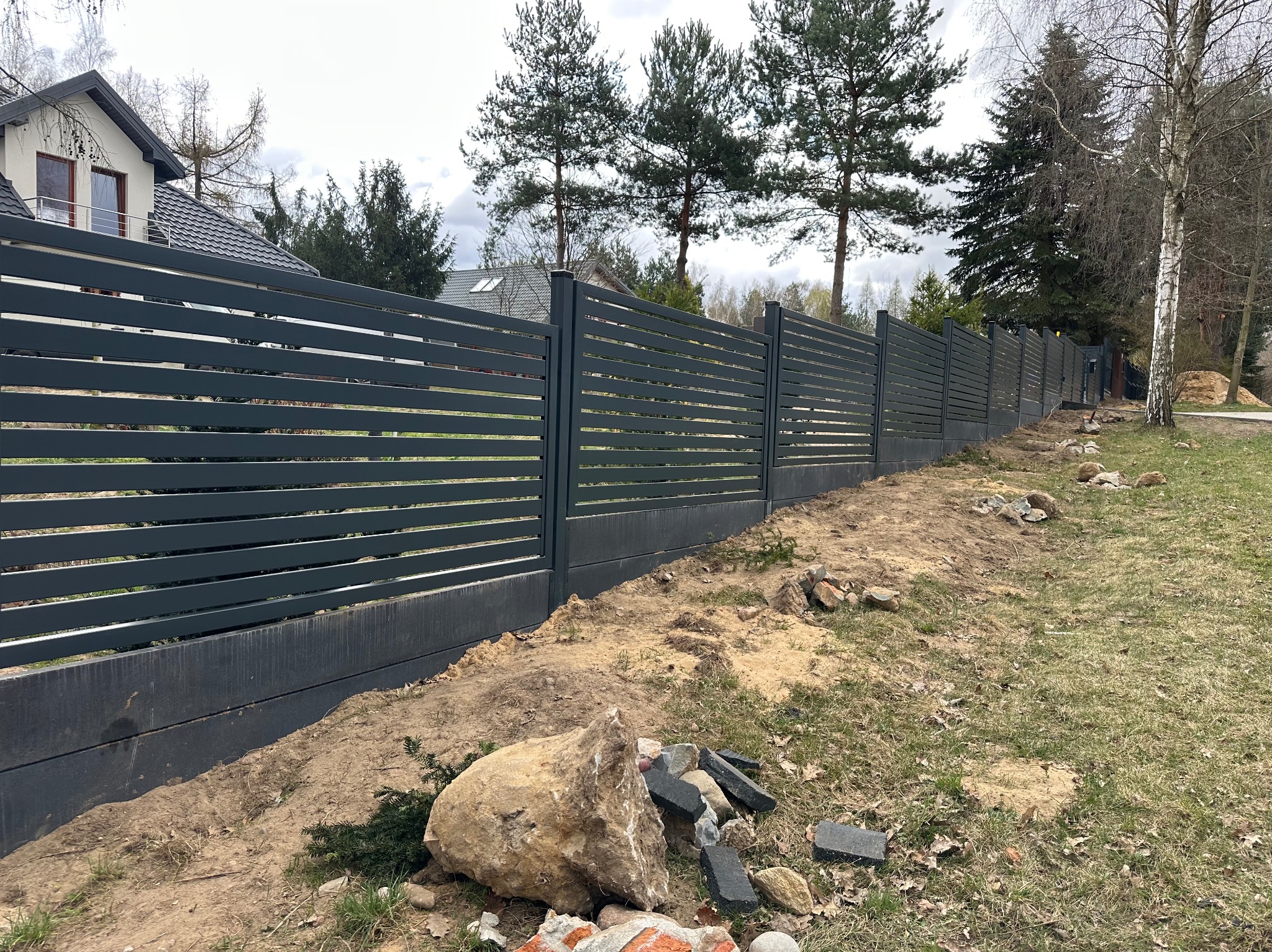 Nowoczesne, poziome ogrodzenie panelowe w kolorze grafitowym, z betonową podmurówką, na tle domu i drzew. Widoczne ślady prac ziemnych.