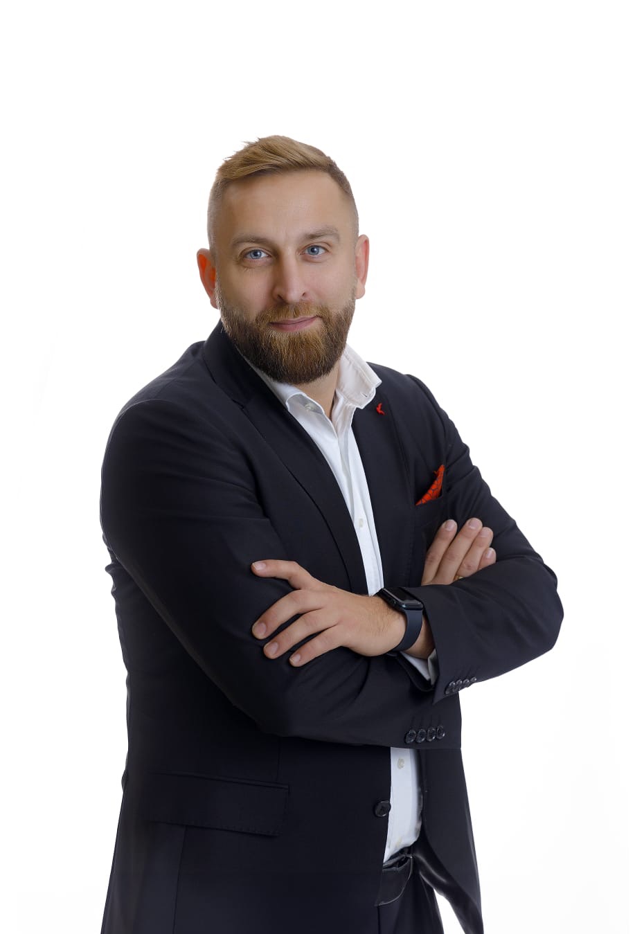 Portret mężczyzny z brodą w czarnym garniturze i białej koszuli, z założonymi rękami na białym tle.