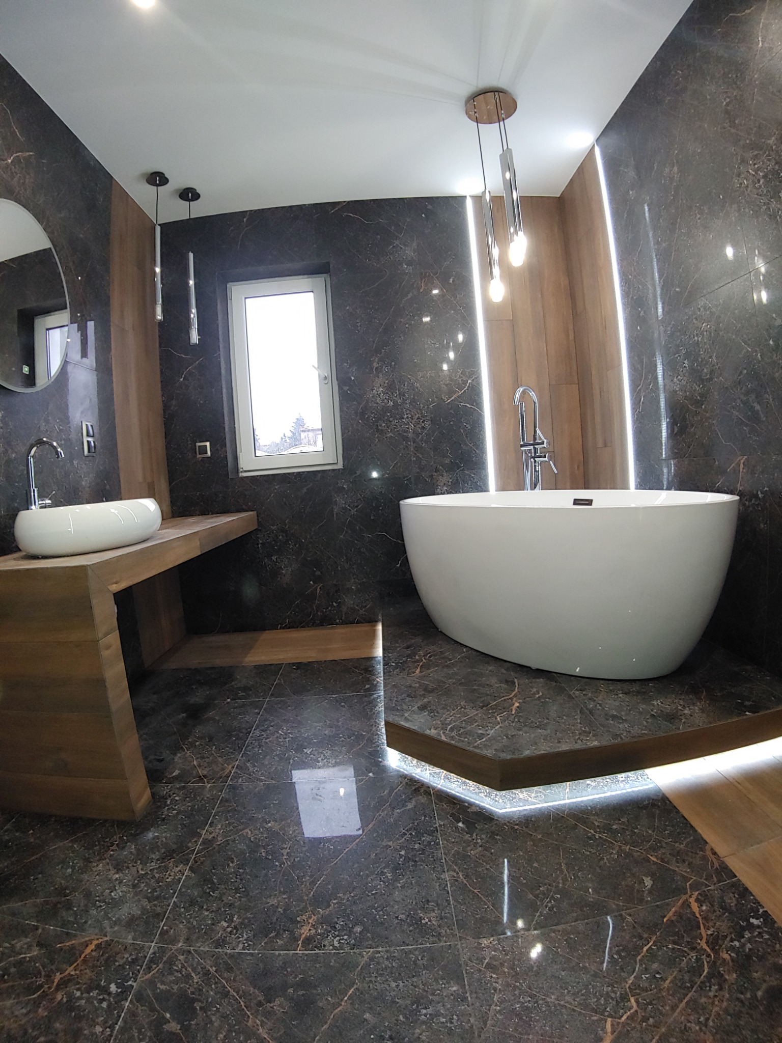 Nowoczesna łazienka z marmurowymi płytkami, wolnostojącą białą wanną, umywalką nablatową na drewnianym blacie i oświetleniem LED.