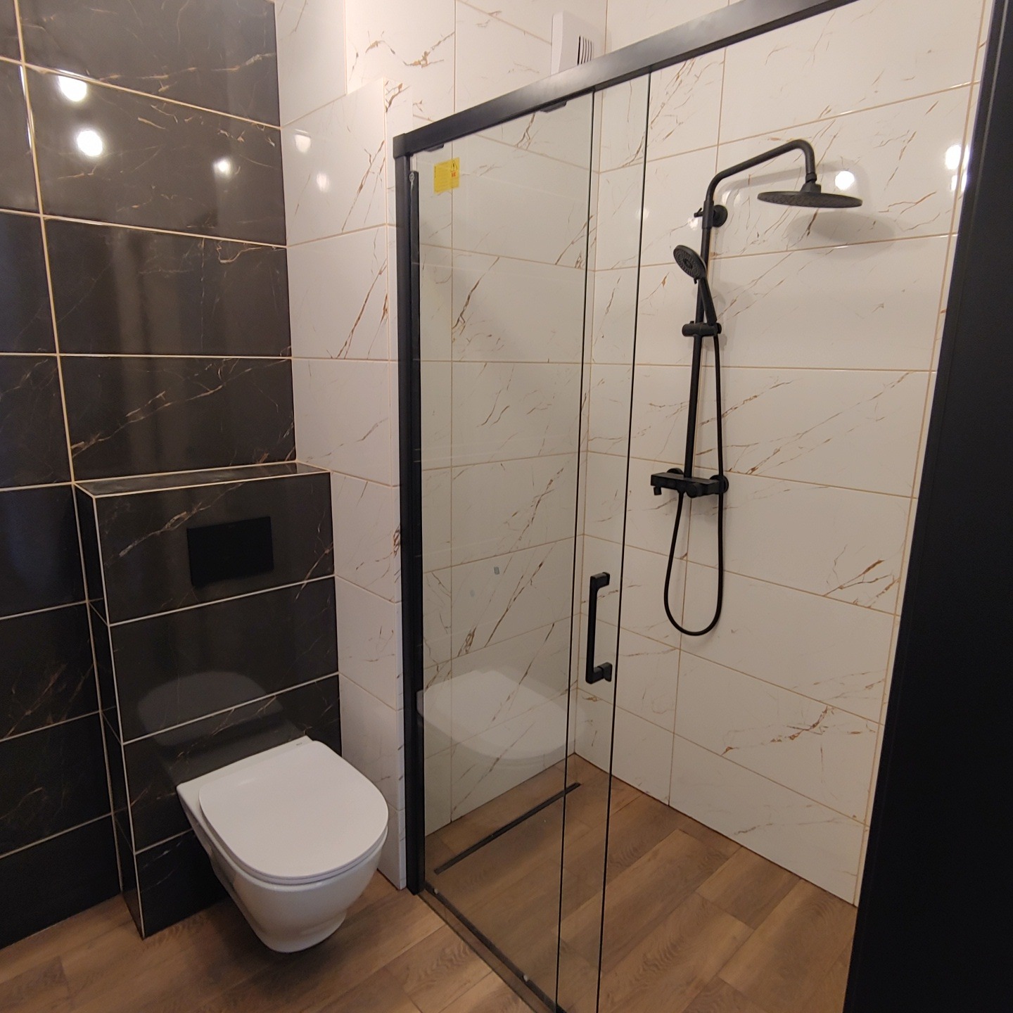 Nowoczesna łazienka z czarnymi akcentami: kabina prysznicowa walk-in z czarną armaturą, wisząca toaleta obudowana ciemnymi płytkami imitującymi marmur, jasne płytki z żyłkami na ścianach...