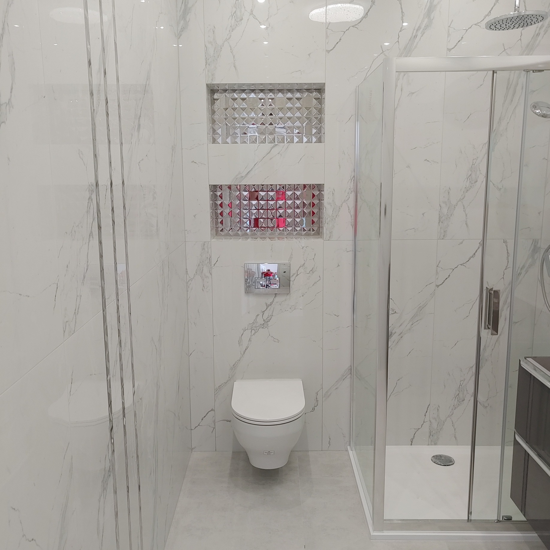 Nowoczesna łazienka z białymi marmurkowymi płytkami, kabiną prysznicową ze szklanymi drzwiami, podwieszaną miską WC i dekoracyjnymi wnękami z mozaiką.