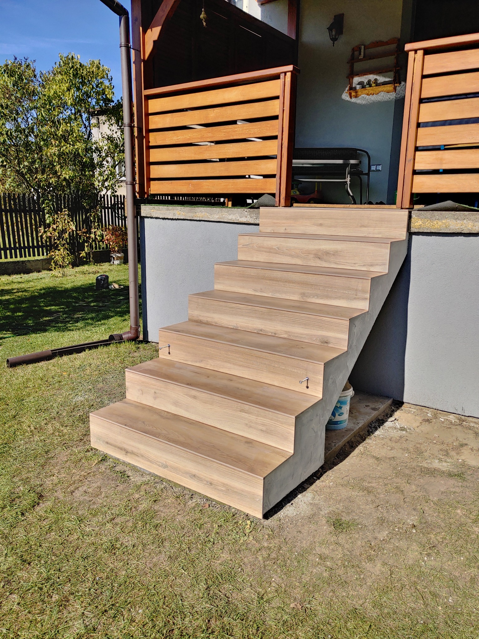 Zewnętrzne schody z drewnopodobną okładziną prowadzące do wejścia domu, z widoczną drewnianą balustradą i rynną w tle.