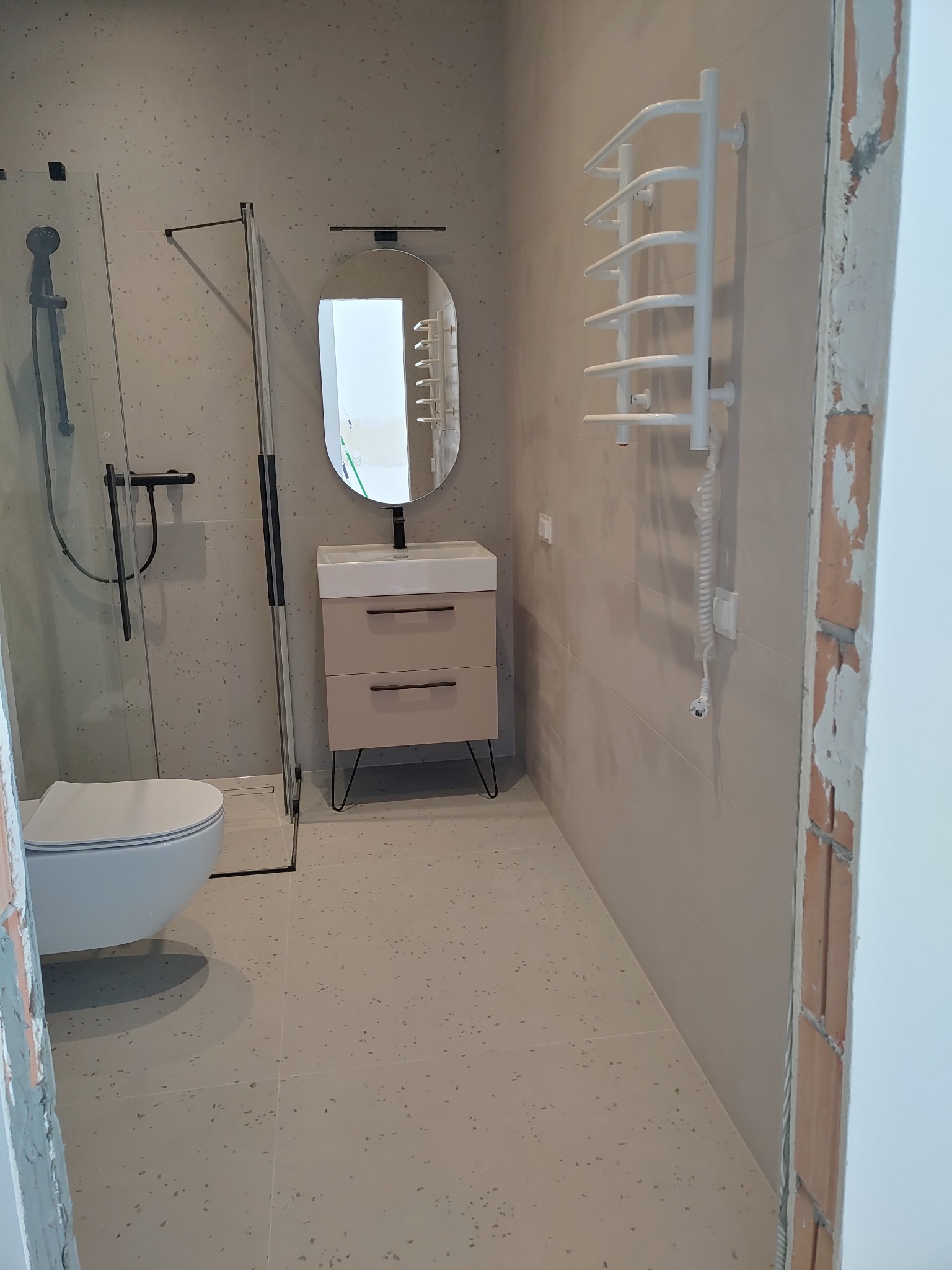 Nowoczesna łazienka z kabiną prysznicową, umywalką z szafką na cienkich nóżkach, grzejnikiem drabinkowym i płytkami imitującymi lastryko na ścianach i podłodze, widoczny fragment surowej ściany...