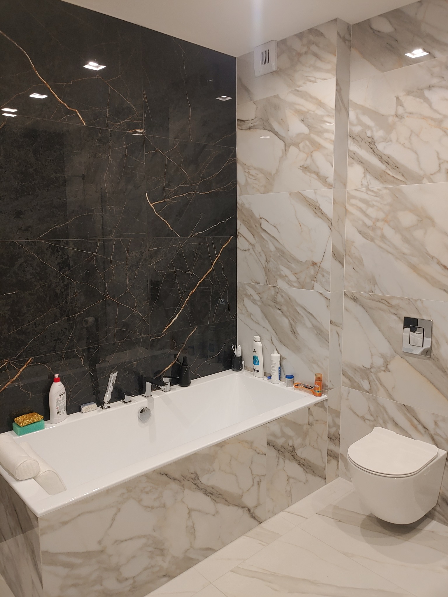 Nowoczesna łazienka z wanną obudowaną marmurowymi płytkami, czarna ściana z marmurowym wzorem, wisząca toaleta, białe płytki podłogowe.