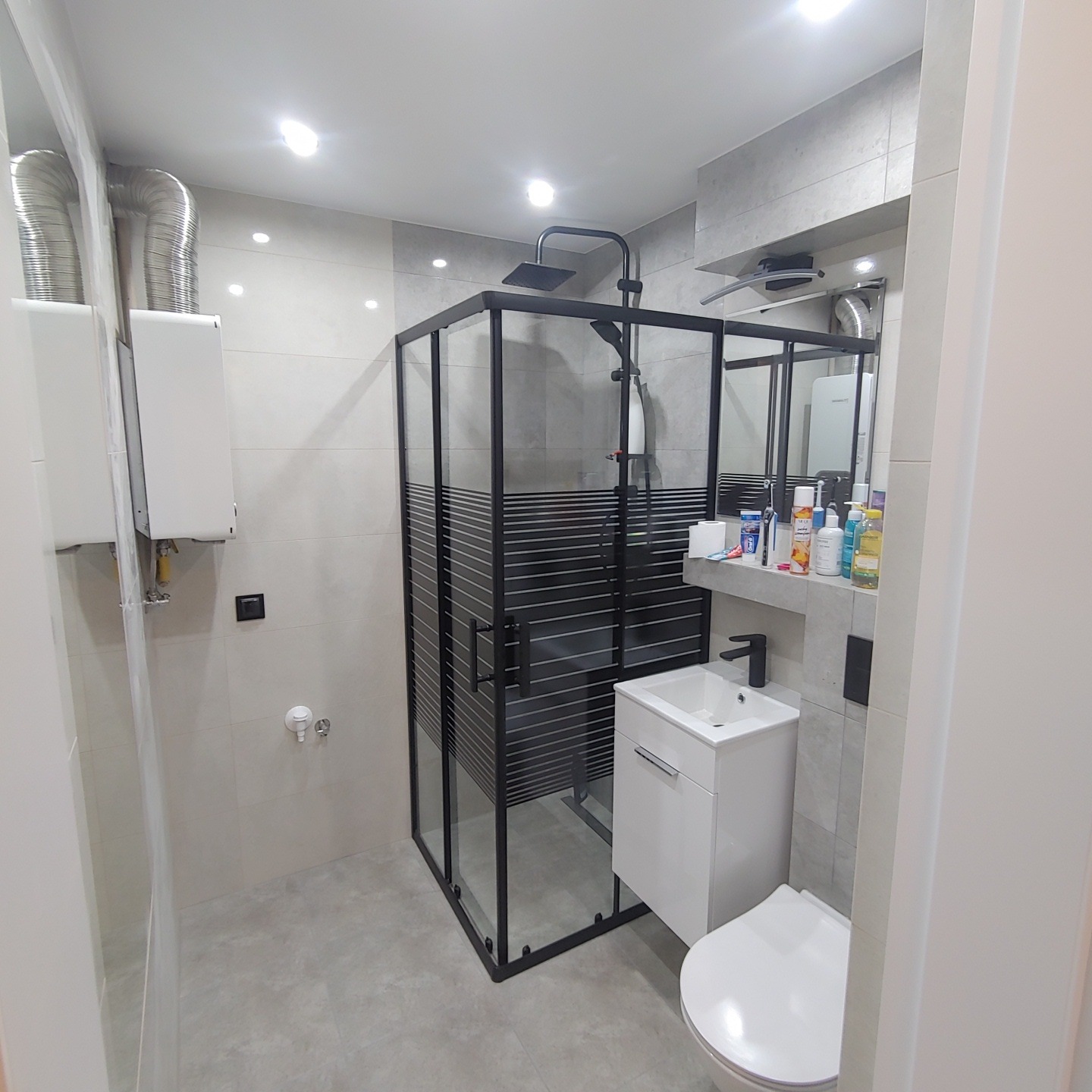 Nowoczesna łazienka z kabiną prysznicową z czarnymi profilami i poziomymi pasami, białą szafką z umywalką i czarną baterią, oraz wiszącą toaletą. Na półce nad umywalką widoczne kosmetyki...