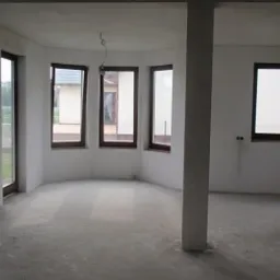 Surowe wnętrze w stanie deweloperskim z widocznymi ścianami po nałożeniu warstwy tynku, świeżo wylana posadzka, duże okna z ciemnymi ramami i kolumna nośna na środku pomieszczenia.