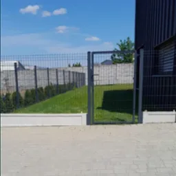 Nowoczesne ogrodzenie panelowe z furtką, widok z zewnątrz na posesję z trawnikiem i tujami, jasne niebo z chmurami.