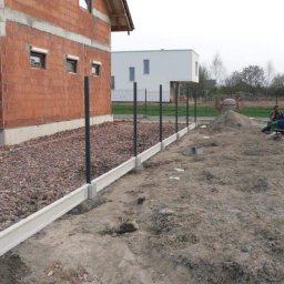 Kamil Nowak Ogrodzenia - Budowa ogrodzenia: betonowa podmurówka z zamontowanymi słupkami na tle surowego budynku z cegły i placu budowy.