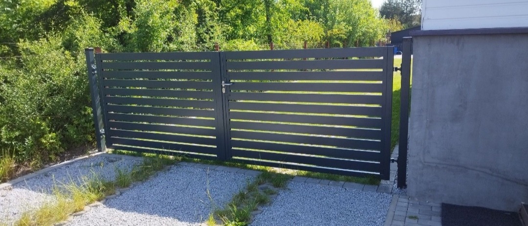 Nowoczesna, antracytowa brama panelowa z poziomymi szczeblami, zamontowana na posesji z szarą nawierzchnią żwirową i zielenią w tle.