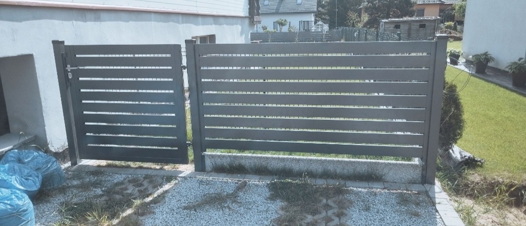 Nowoczesne, poziome ogrodzenie panelowe w kolorze antracytowym z furtką, zamontowane na posesji z widocznym trawnikiem i domem w tle.