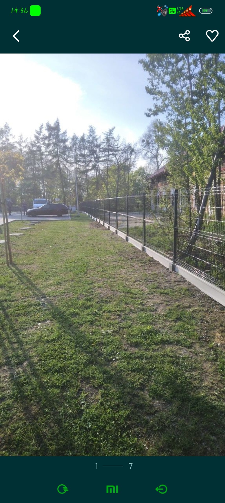 Długi, szary panelowy płot z siatki na betonowej podmurówce, oddzielający trawnik od drzew i budynków w tle.