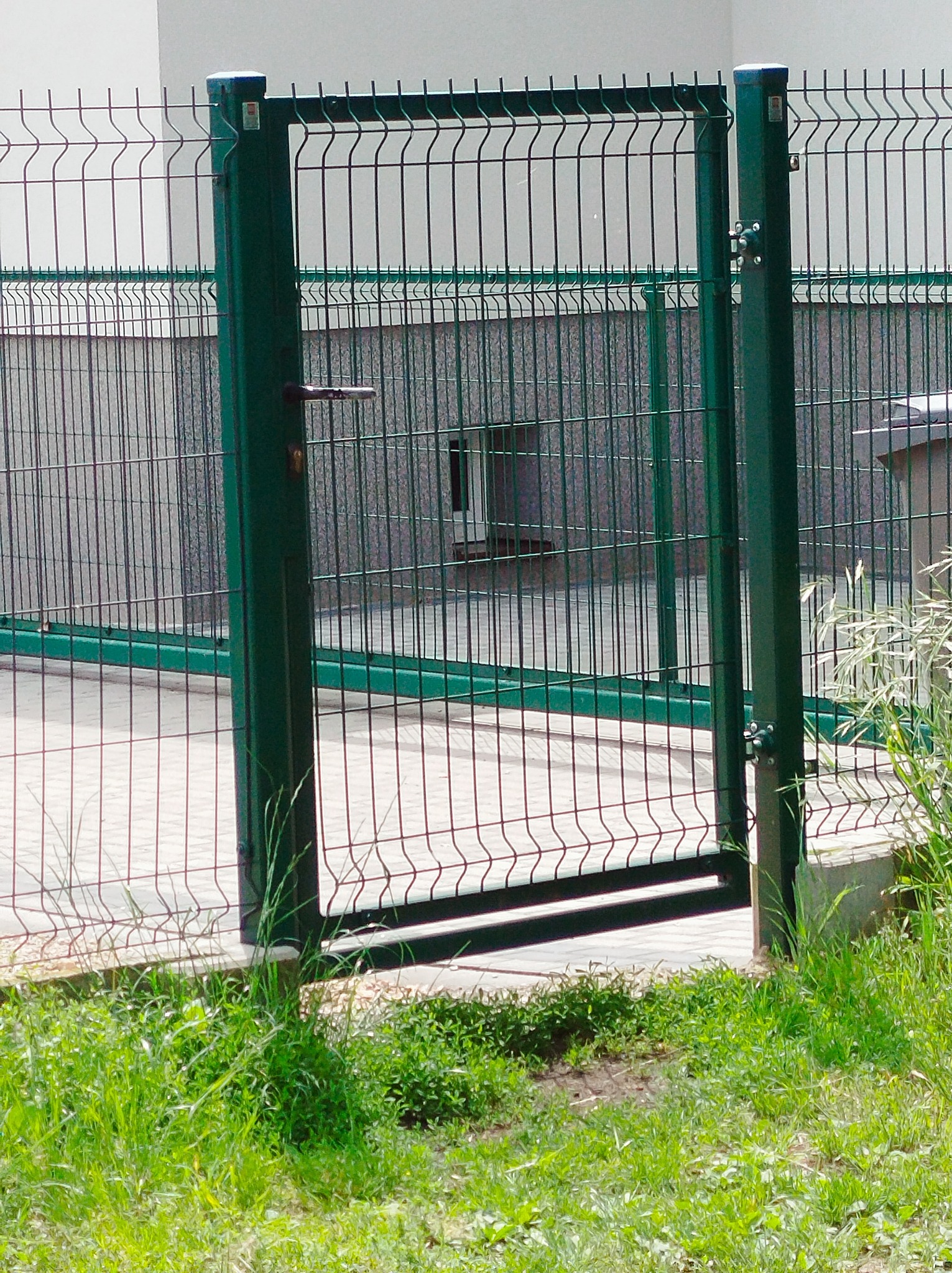 Otwarta furtka w zielonym ogrodzeniu panelowym z widoczną klamką i zawiasami, na tle jasnego budynku i kostki brukowej, z fragmentem trawy w dolnej części kadru.