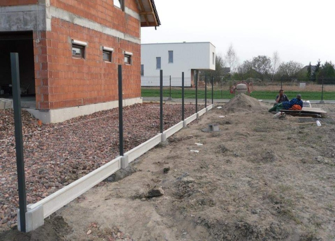Budowa ogrodzenia: betonowa podmurówka z zamontowanymi słupkami na tle surowego budynku z cegły i placu budowy.