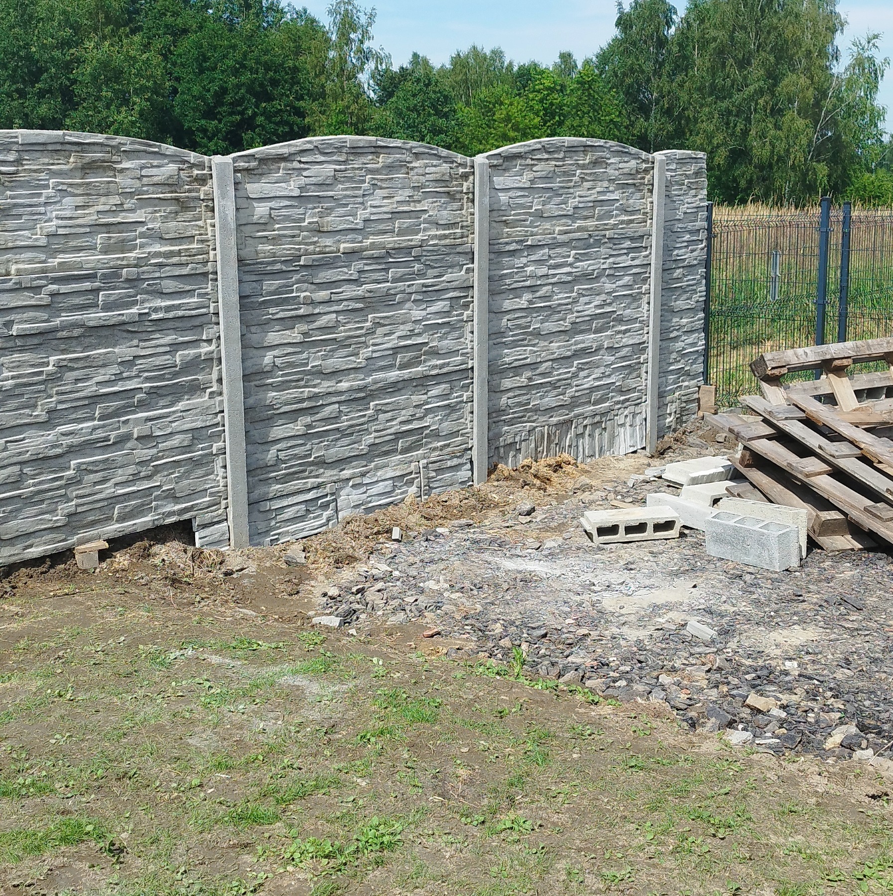 Fragment nowo postawionego, szarego ogrodzenia z paneli imitujących kamień, widoczne betonowe słupki, a w tle teren z resztkami materiałów budowlanych, paletami i zielenią.