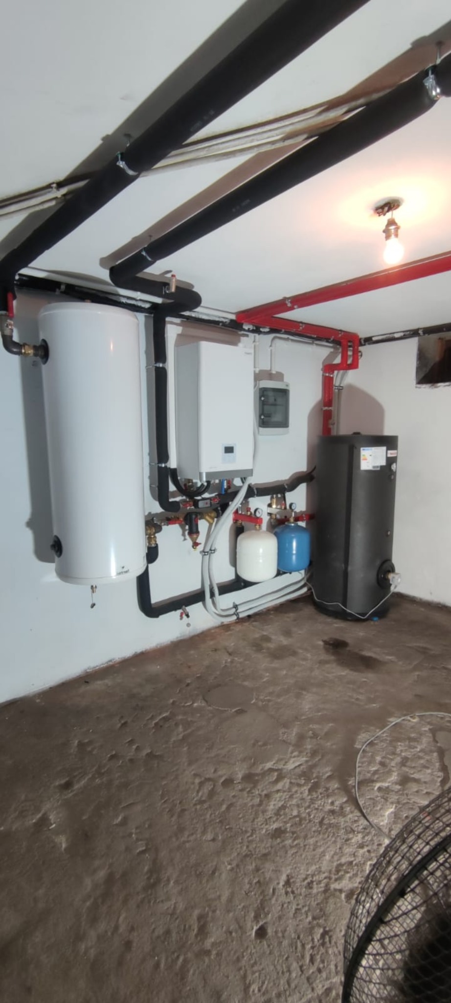 Zakończona instalacja grzewcza w piwnicy: biały zasobnik ciepłej wody, piec gazowy, czarny bojler, rury w izolacji termicznej, zbiorniki wyrównawcze. Szare ściany i podłoga.