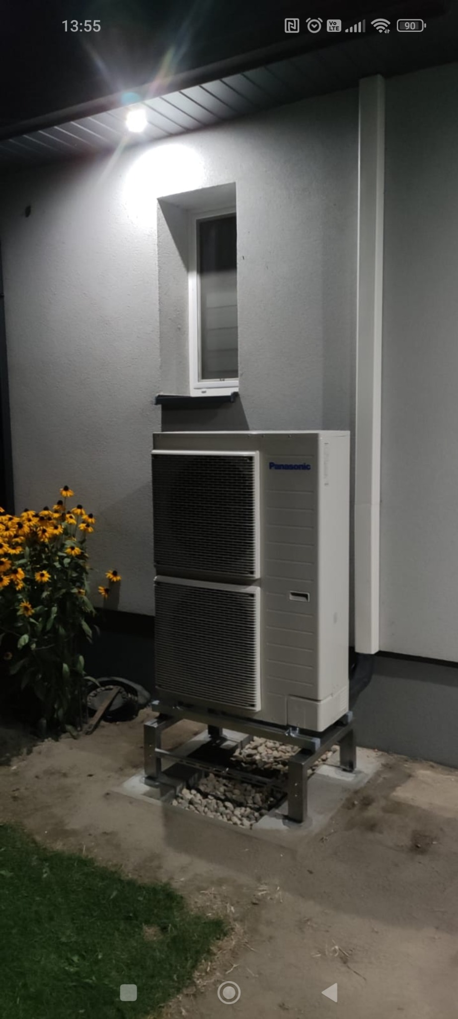 Zewnętrzna jednostka pompy ciepła Panasonic na metalowej konstrukcji, obok kwitnące rudbekie, zamontowana przy ścianie budynku z małym oknem.