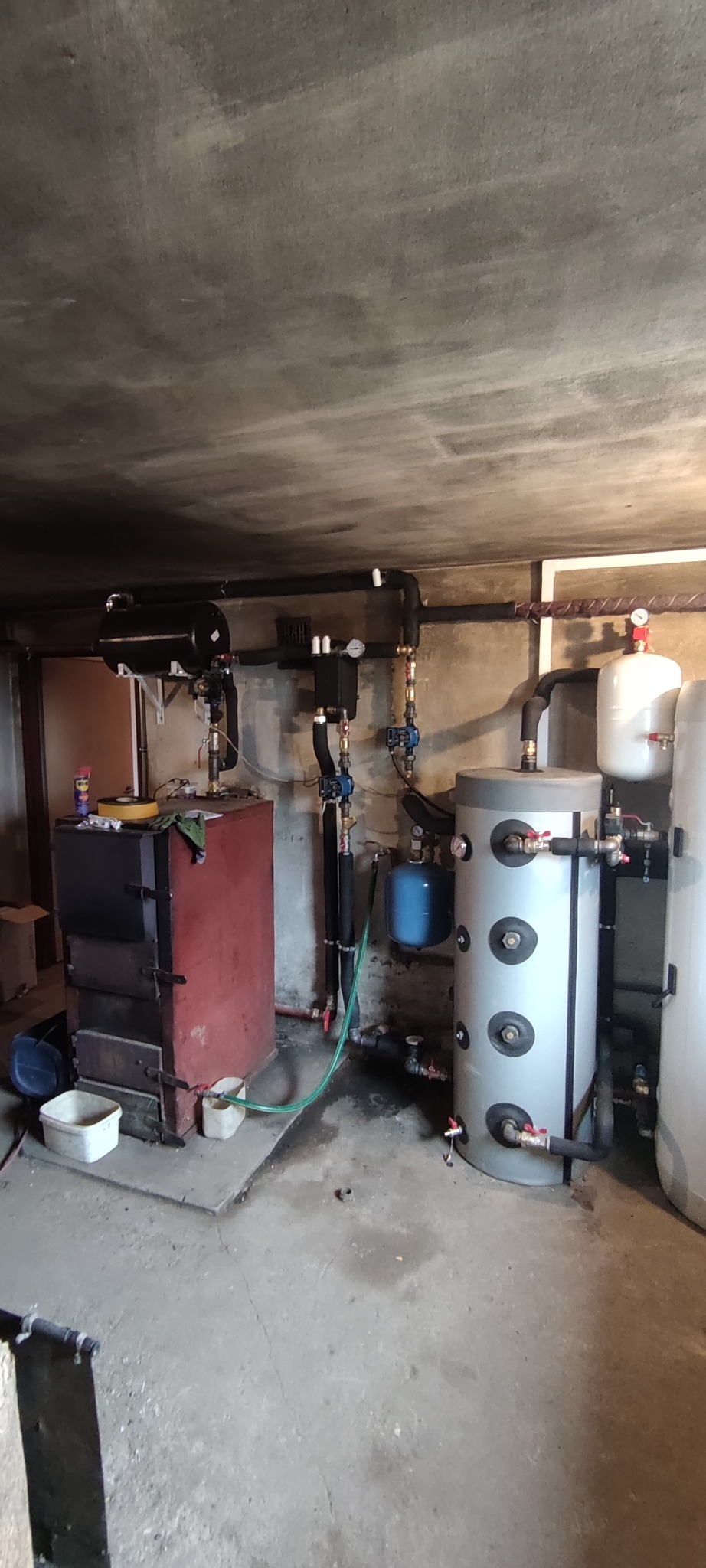 Kotłownia z piecem na paliwo stałe, zbiornikami akumulacyjnymi i instalacją hydrauliczną. Widoczne rury, zawory i osprzęt. Surowy beton na ścianach i suficie.