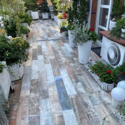 Igor Martsynkovskyi - Długi, wąski balkon z podłogą wyłożoną płytkami imitującymi drewno w odcieniach szarości, brązu i błękitu, otoczony różnorodną zielenią w białych donicach i skrzynkach, z widocznym klimatyzatorem...