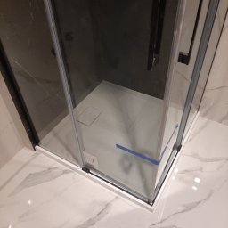 Igor Martsynkovskyi - Szklana kabina prysznicowa z czarnymi uchwytami, zabezpieczona niebieską taśmą na dole, osadzona na marmurowej podłodze z widocznym odpływem liniowym.