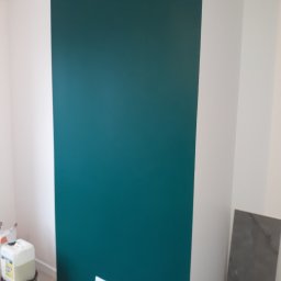 Igor Martsynkovskyi - Świeżo pomalowana, gładka ściana w odcieniu głębokiej zieleni z widocznymi gniazdkami elektrycznymi, obok biała ściana i wiadro z farbą w tle.