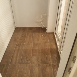 Igor Martsynkovskyi - Balkon po remoncie z brązowymi płytkami imitującymi drewno, białe ściany, widoczne niepodłączone kable i roleta okienna.