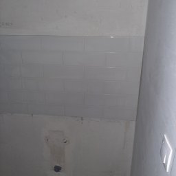 Igor Martsynkovskyi - Ściana łazienki w trakcie układania płytek, widoczne przygotowane otwory na armaturę, częściowo położone białe, prostokątne płytki na szarej, surowej ścianie.