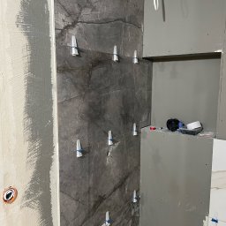 Igor Martsynkovskyi - Montaż płytek imitujących marmur na ścianie w łazience z użyciem klipsów poziomujących, widoczna surowa ściana z gładzią oraz płyta karton gips z wnęką, w której leżą akcesoria glazurnicze.