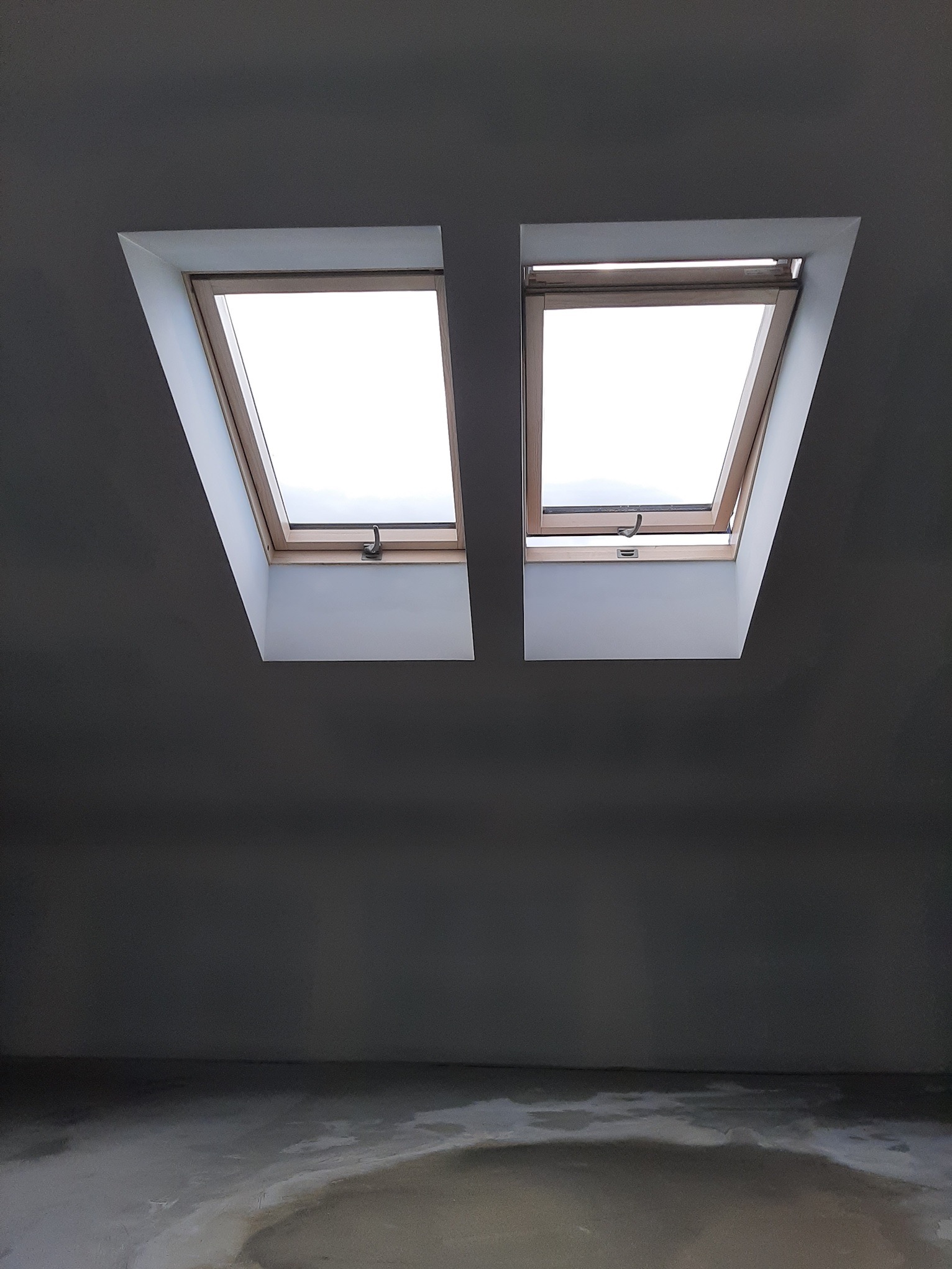 Dwa okna dachowe w szarym pomieszczeniu ze ścianami pomalowanymi na szaro i surową podłogą z widocznymi śladami wilgoci.