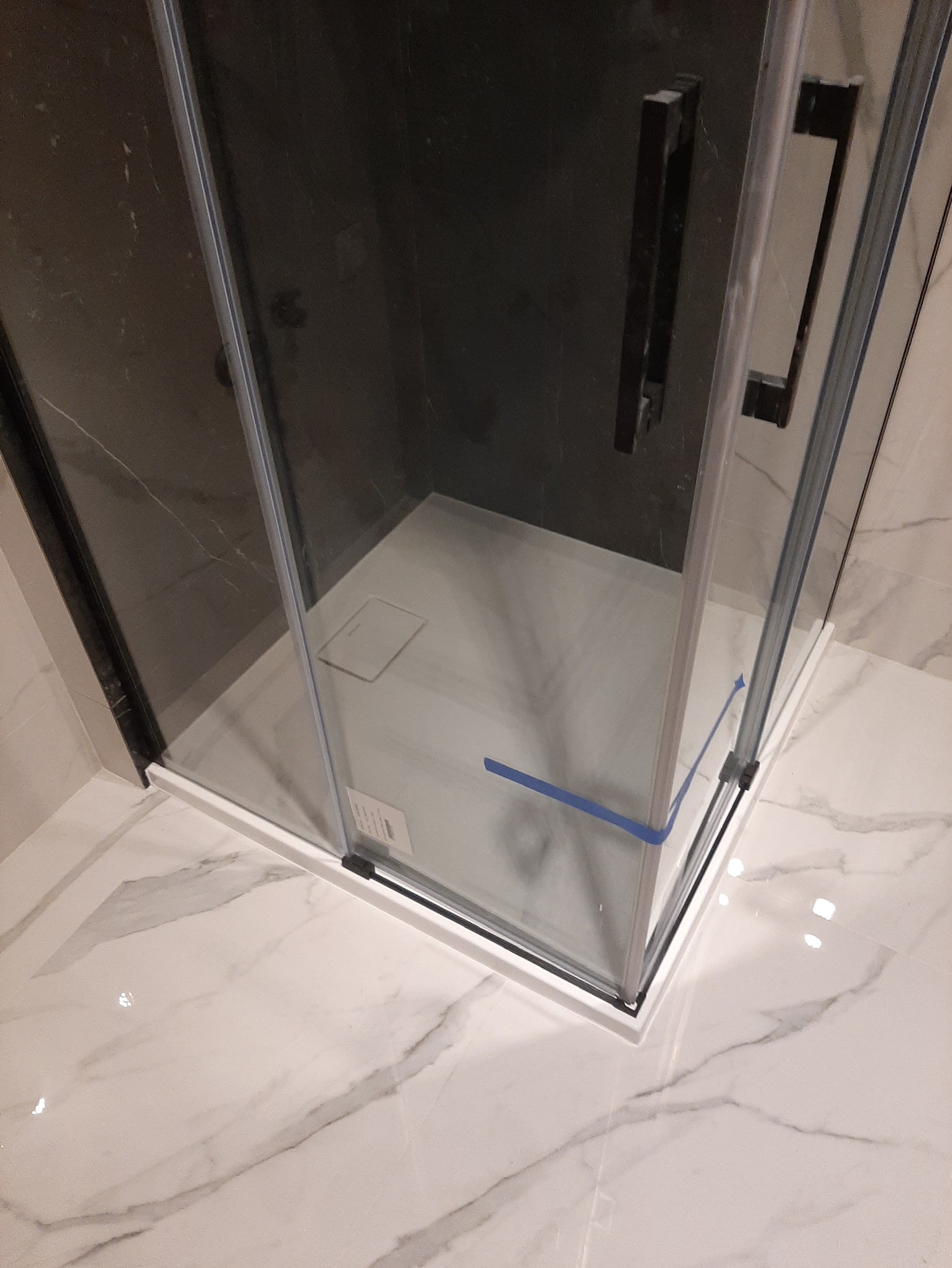 Szklana kabina prysznicowa z czarnymi uchwytami, zabezpieczona niebieską taśmą na dole, osadzona na marmurowej podłodze z widocznym odpływem liniowym.