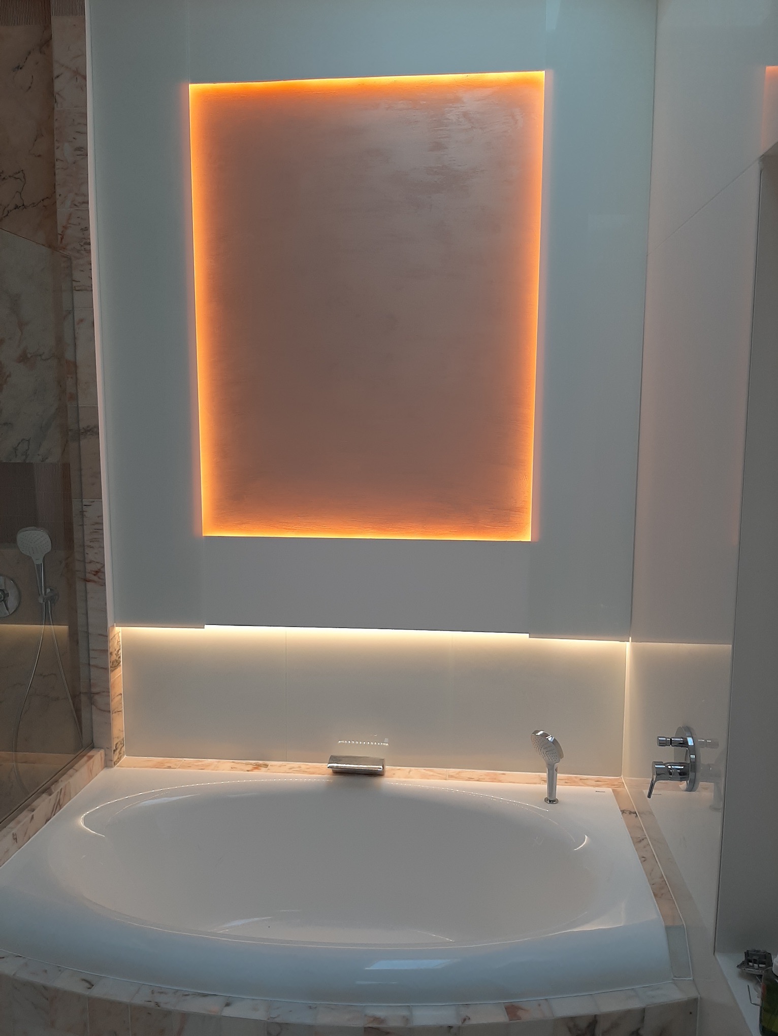 Nowoczesna łazienka z białą wanną obudowaną marmurem, podświetlonym panelem ściennym i minimalistyczną armaturą.