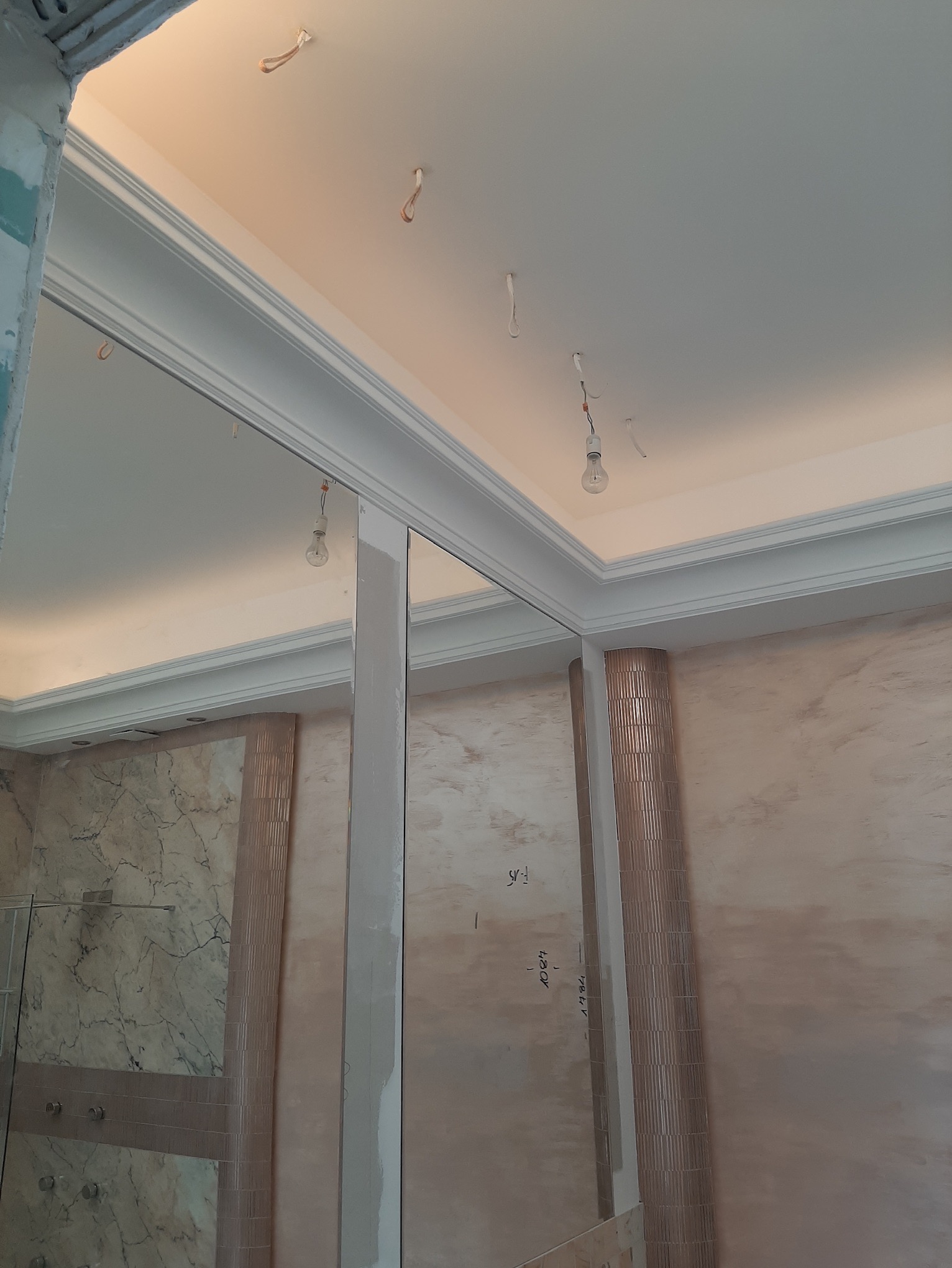 Fragment łazienki w trakcie prac: widoczne niedokończone ściany z dekoracyjnymi paskami, sufit z listwami i wiszącymi przewodami elektrycznymi, oraz częściowo zamontowana kabina prysznicowa.