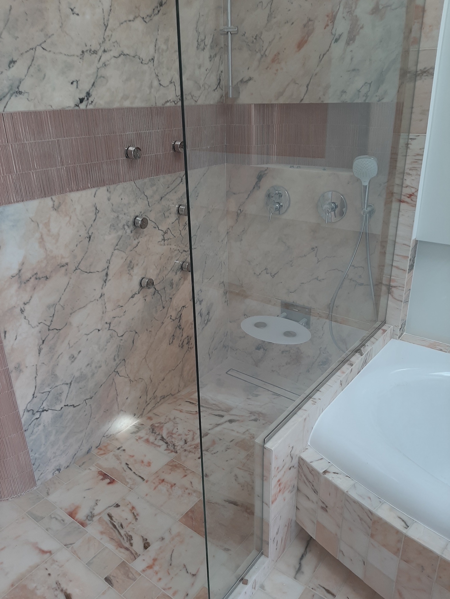 Nowoczesna kabina prysznicowa z marmurowymi ścianami i podłogą w odcieniach bieli i różu, szklana ściana oddzielająca od wanny, widoczne dysze prysznicowe i bateria.
