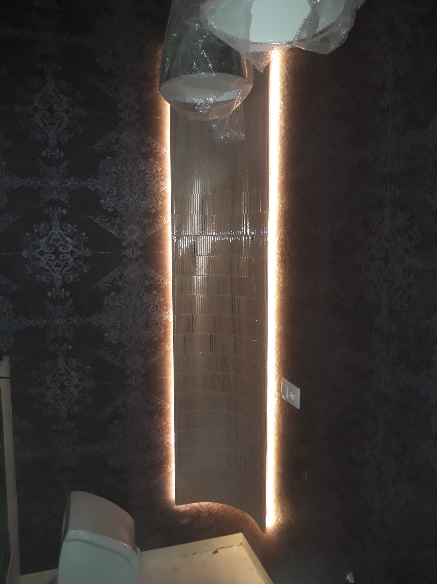 Fragment łazienki w trakcie prac: podświetlona ściana z dekoracyjnymi płytkami, widoczna część toalety jeszcze w folii ochronnej i ściana z ciemną tapetą w ornamenty.