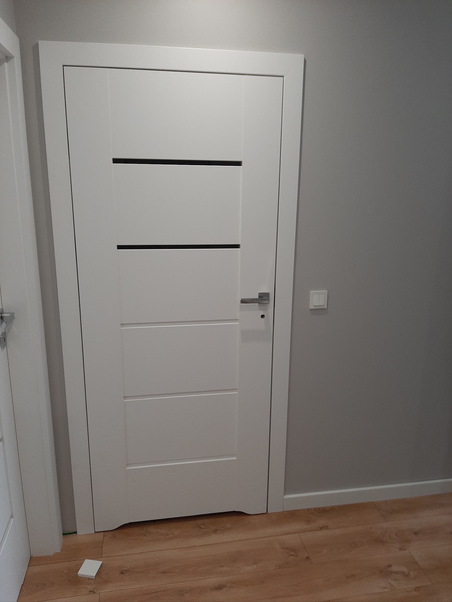 Białe, nowoczesne drzwi wewnętrzne z poziomymi wstawkami na tle szarej ściany i drewnopodobnej podłogi, widoczny fragment białej listwy przypodłogowej i włącznik światła.