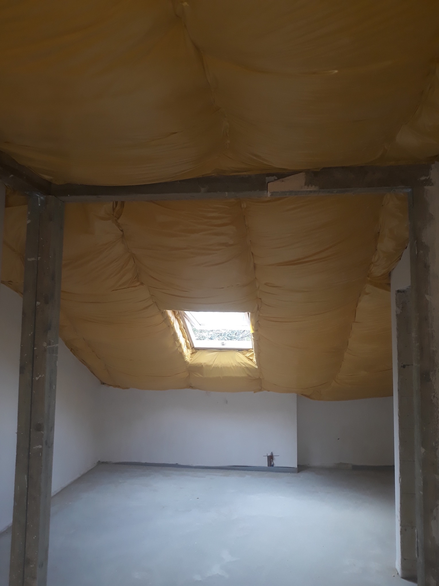 Wnętrze w trakcie prac, widoczna izolacja stropu folią w kolorze żółtym, betonowa wylewka na podłodze, surowe ściany i framugi drzwi.