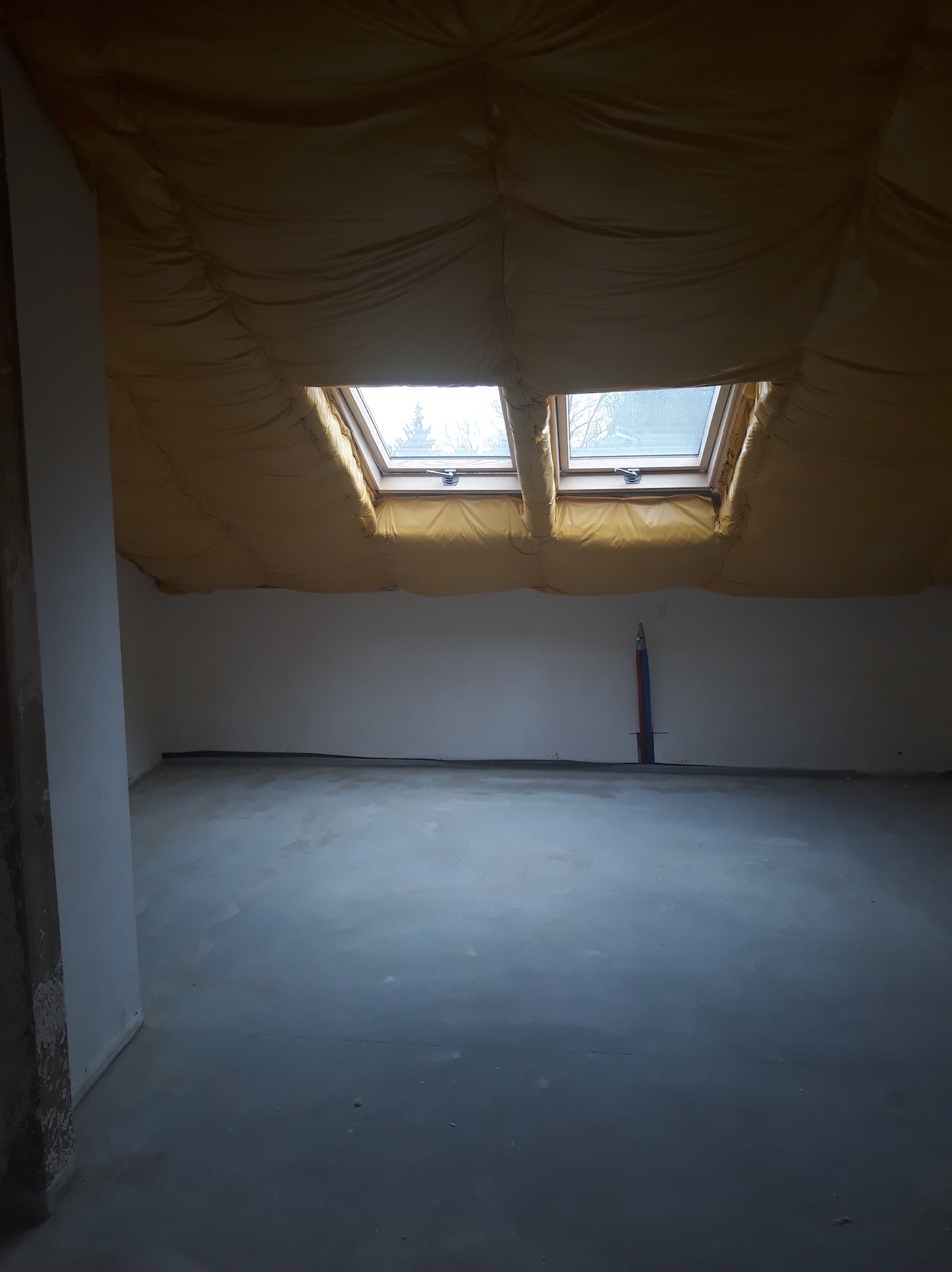 Poddasze w trakcie prac przygotowawczych: widoczne okna dachowe, izolacja stropu folią, surowa ściana i wylewka podłogowa, pion instalacyjny na ścianie.