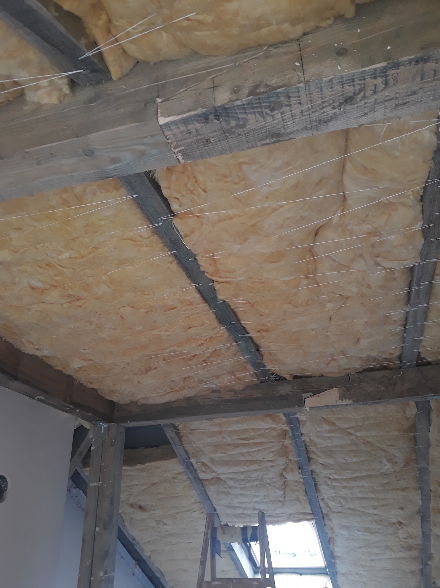 Ocieplenie poddasza wełną mineralną, widoczne drewniane belki konstrukcyjne i sznurki podtrzymujące izolację przed montażem płyt kartonowo-gipsowych.