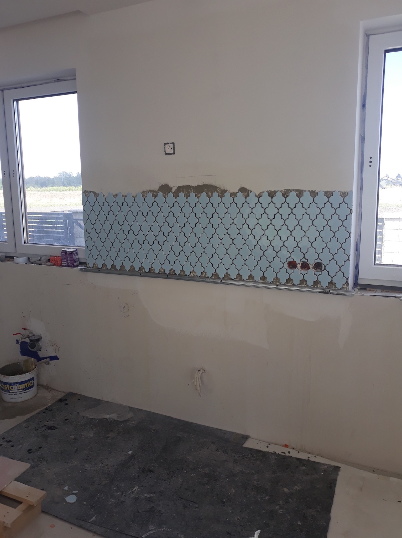 Ściana w trakcie układania mozaiki marokańskiej w odcieniach błękitu, widoczne puszki elektryczne i poziomica, okno z widokiem na zewnątrz.