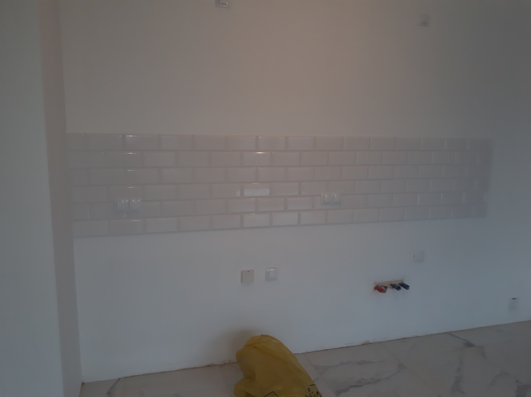 Ściana w trakcie układania białych, błyszczących płytek w kształcie cegiełek, widoczne przygotowane instalacje wodno-kanalizacyjne oraz worki z materiałami budowlanymi na podłodze.