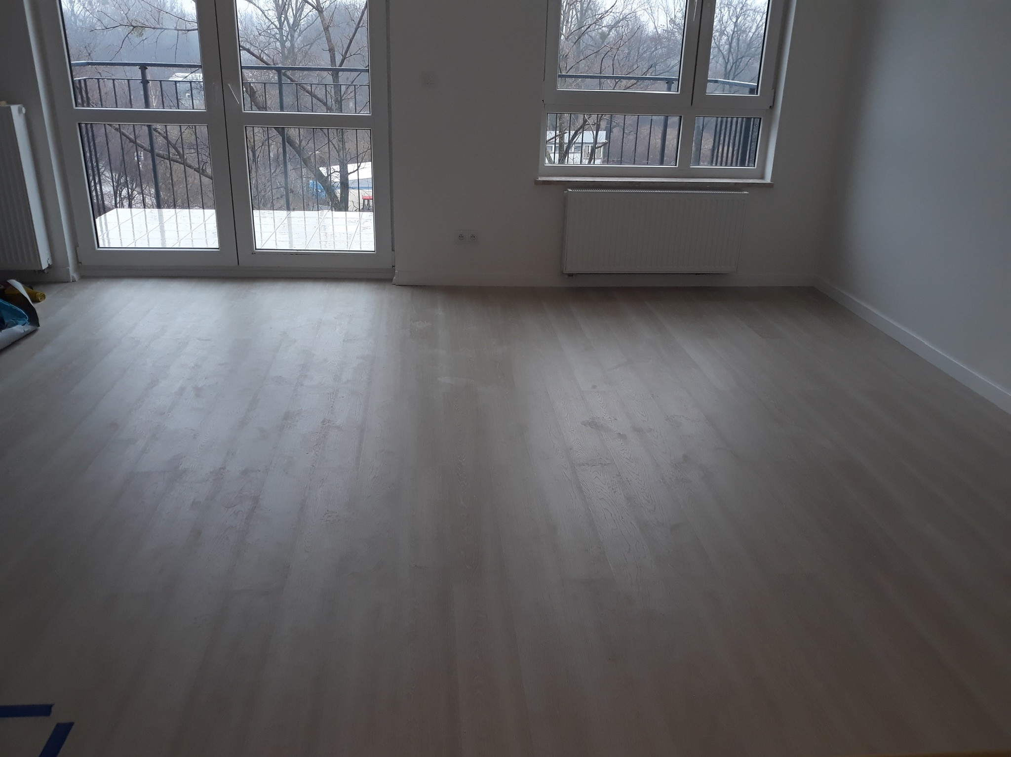 Jasna, pusta przestrzeń z nową drewnianą podłogą, białe ściany, okno z widokiem na drzewa bez liści i balkon z czarną balustradą, grzejnik pod oknem.