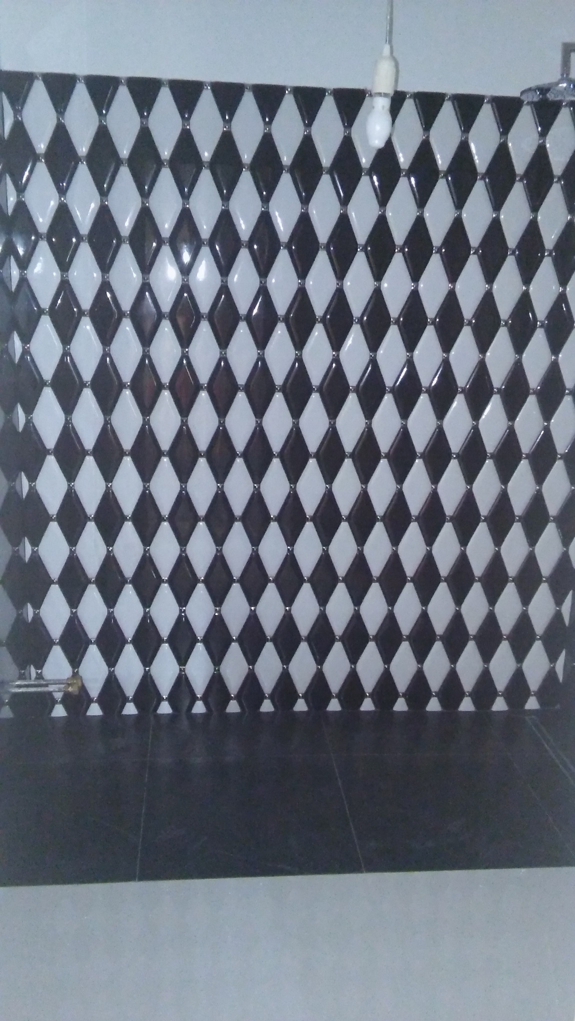 Ściana wyłożona błyszczącymi, romboidalnymi płytkami w szachownicę czarno-białą, z ciemną podłogą i widoczną żarówką na kablu u góry.