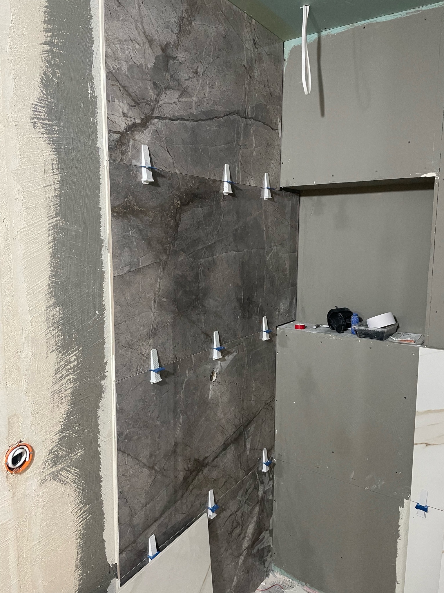 Montaż płytek imitujących marmur na ścianie w łazience z użyciem klipsów poziomujących, widoczna surowa ściana z gładzią oraz płyta karton gips z wnęką, w której leżą akcesoria glazurnicze.