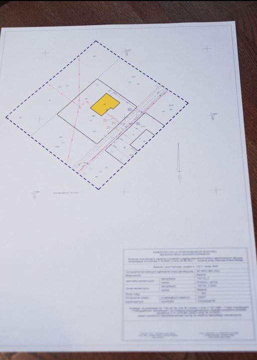 Plan zagospodarowania przestrzennego działki z zaznaczonym budynkiem w kolorze żółtym i naniesionymi elementami infrastruktury, umieszczony na białym arkuszu papieru leżącym na drewnianym podłożu.