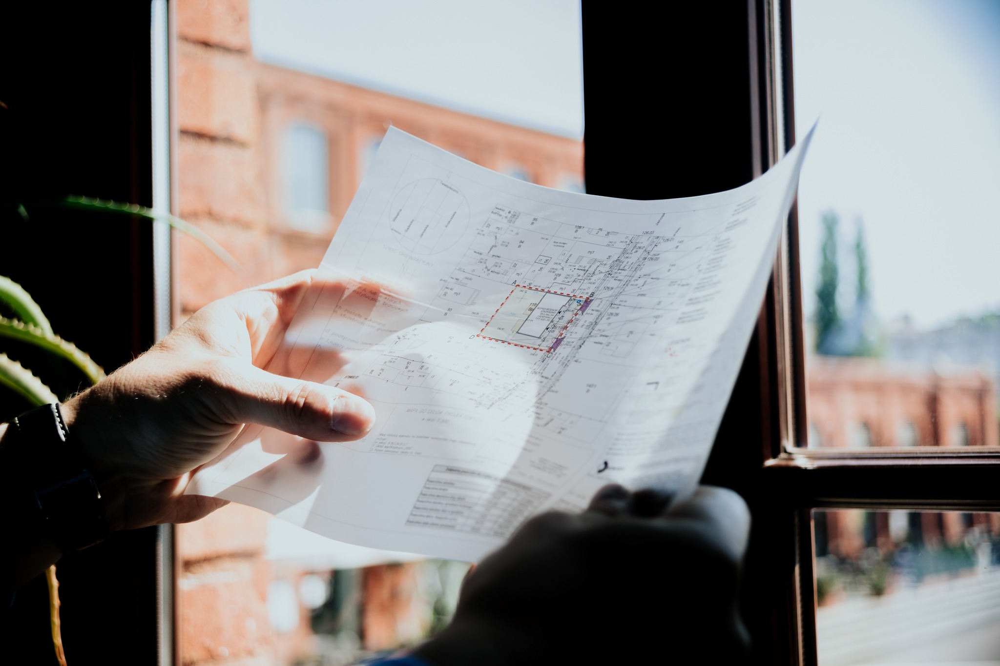 Ręce trzymające plan działki z zaznaczonymi granicami budynku, widoczne przez okno z ceglanym budynkiem w tle.