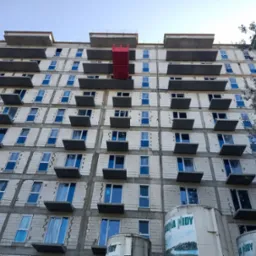 Wielopiętrowy budynek w stanie surowym z czerwonym podnośnikiem na elewacji, widok z dołu, okna z niebieskimi ramami, zbiorniki z napisem Nidy w dolnej części kadru.