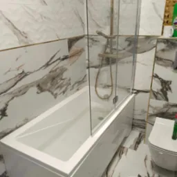 Biała wanna z parawanem nawannowym i złotą armaturą na tle marmurowych płytek w łazience. Widoczny fragment toalety i środki czystości.