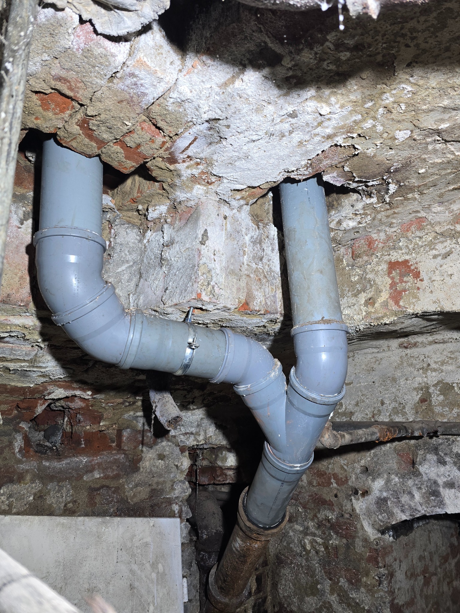 Szare rury kanalizacyjne z łącznikami w starej, ceglanej piwnicy. Widoczne ślady wilgoci i korozji na ścianach i rurach. Rury przymocowane metalową obejmą.